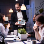 Entrepreneur businessman showing company strategy using tablet for corporate presentation working overtime in meeting office room late at night. Diverse multi-ethnic teamwork brainstorming ideas.